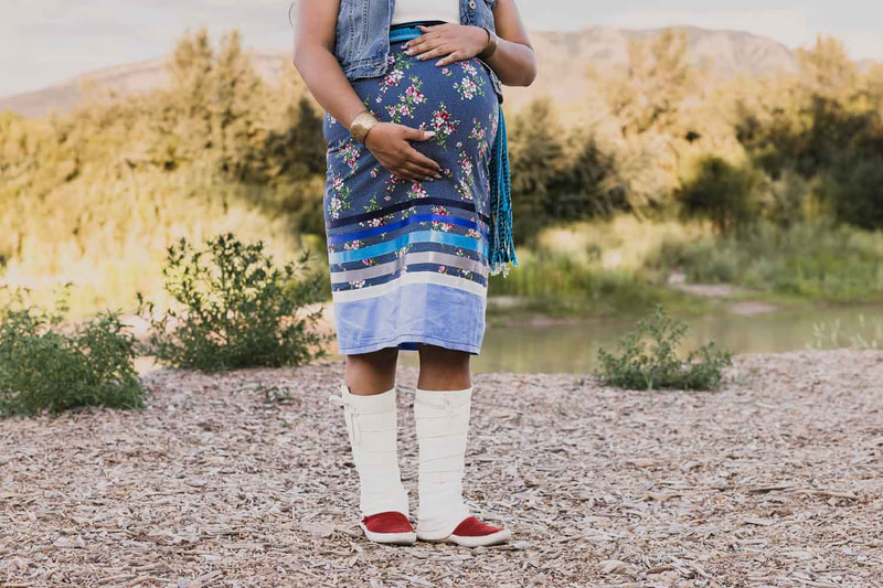 Pregnant mother stands in front of river in ribbon skirt.